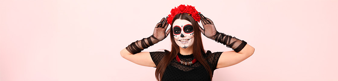 Foto de una mujer sonriente, maquillada de Catrina, poniéndose su corona de flores de papel frente a un fondo rosado